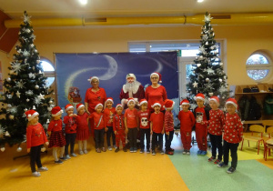 Grupa dzieci stoi z panią dyrektor Marią Królikowską i panią Arletą Kalinowską, w tle dekoracja świąteczna, choinki.